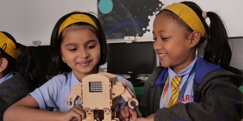 Two schoolgirls in uniforms, playing with a humanoid robot in a classroom 