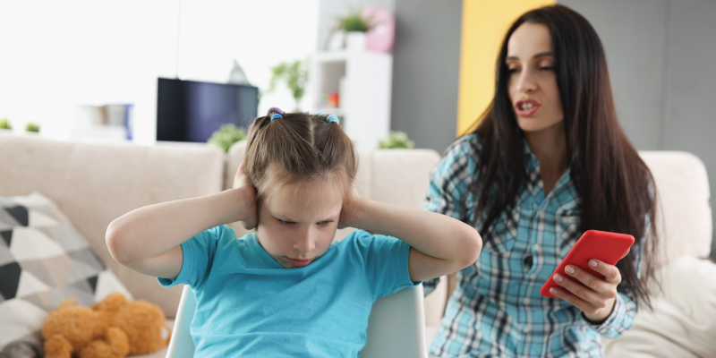 A young girl covers her ears as her mom scolds her for using her mobile during homework, questioning the tech's fit for her age