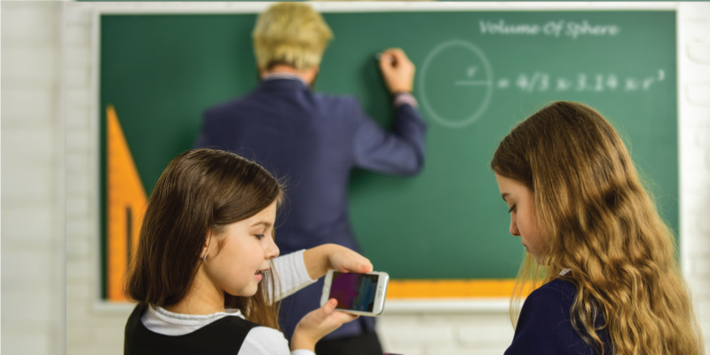 Two young girls use a smartphone in class while the teacher writes on the blackboard, showcasing how technology can distract students