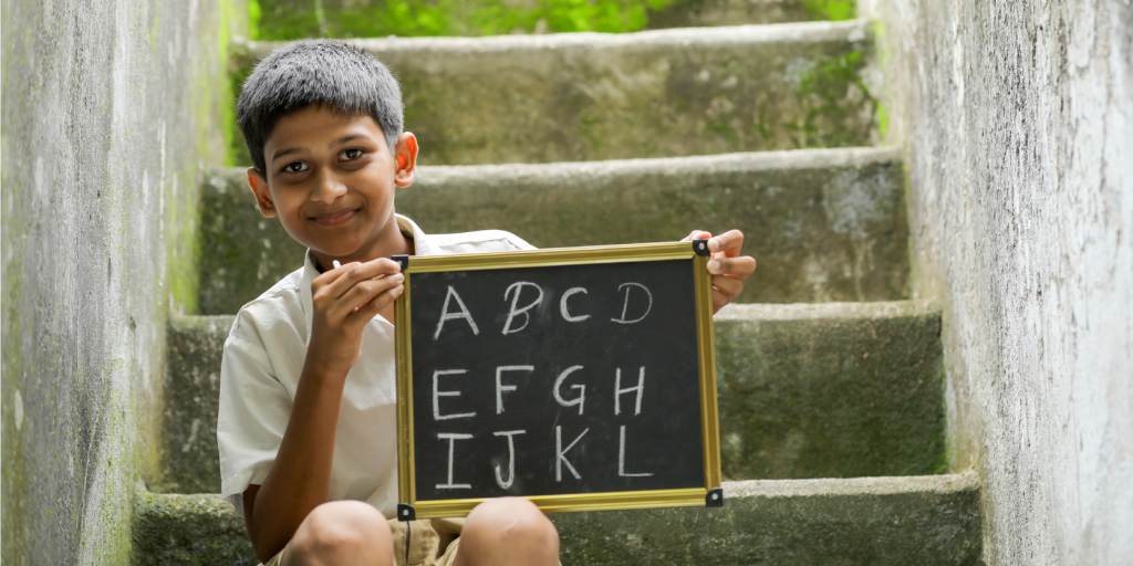 A young boy proudly displays a chalkboard with the alphabet, symbolizing educational advancement under Sarva Shiksha Abhiyan