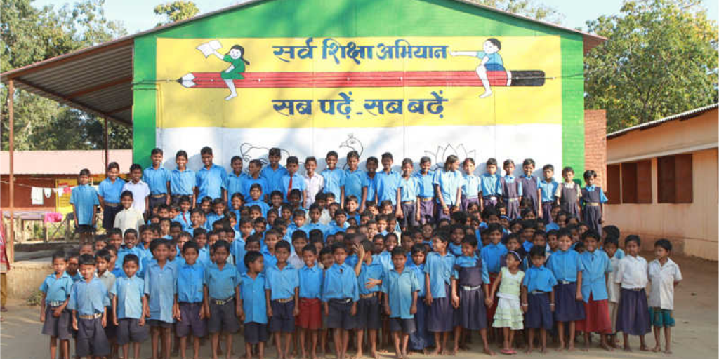 A large group of government school students gathered under the vibrant educational mural of Sarva Siksha Abhiyan