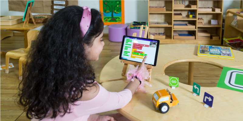 A young girl is using PictoBlox AI and Quarky DIY AI and Robotics kit on a tablet in a classroom setting