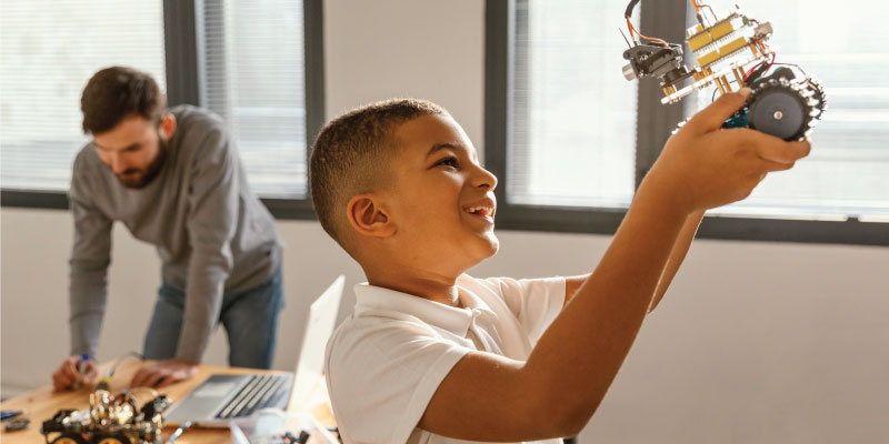 A young boy joyfully holds a robot he built while an adult works in the background, embodying 21st century STEAM education